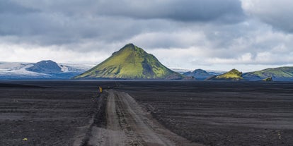 A black volcanic dirt road stretches toward the base of Maelifell Volcano, creating a sharp contrast with the mountain’s vibrant green slopes.