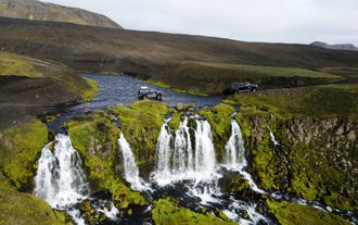 Two super jeeps cross a river that cascades into a small waterfall during an off-road adventure in the Highlands of Iceland.