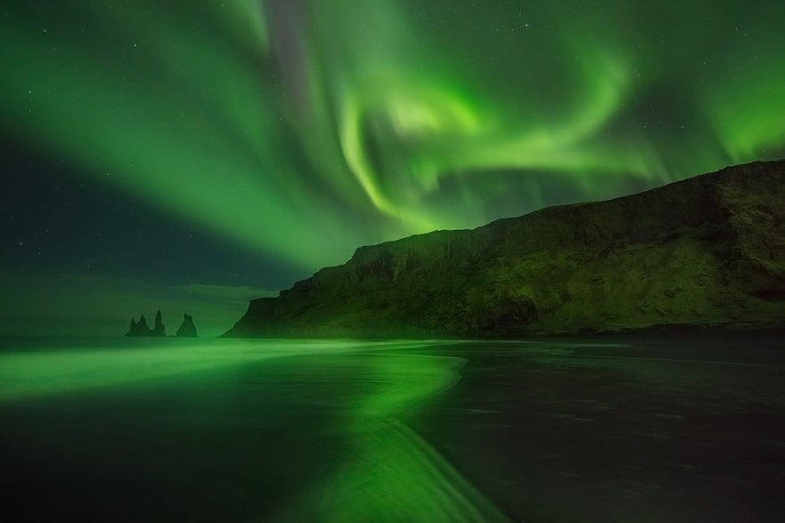 Aurora shines over the black sand beach near Vik, with Reynisdrangar Sea Stacks visible in the distance.