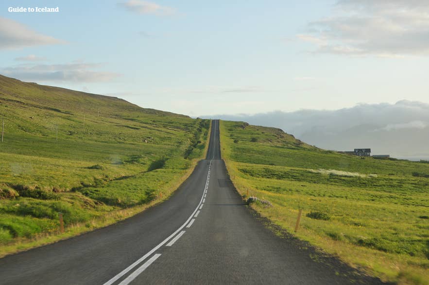 Empty two-lane paved road stretching through green countryside toward distant hills in Iceland.