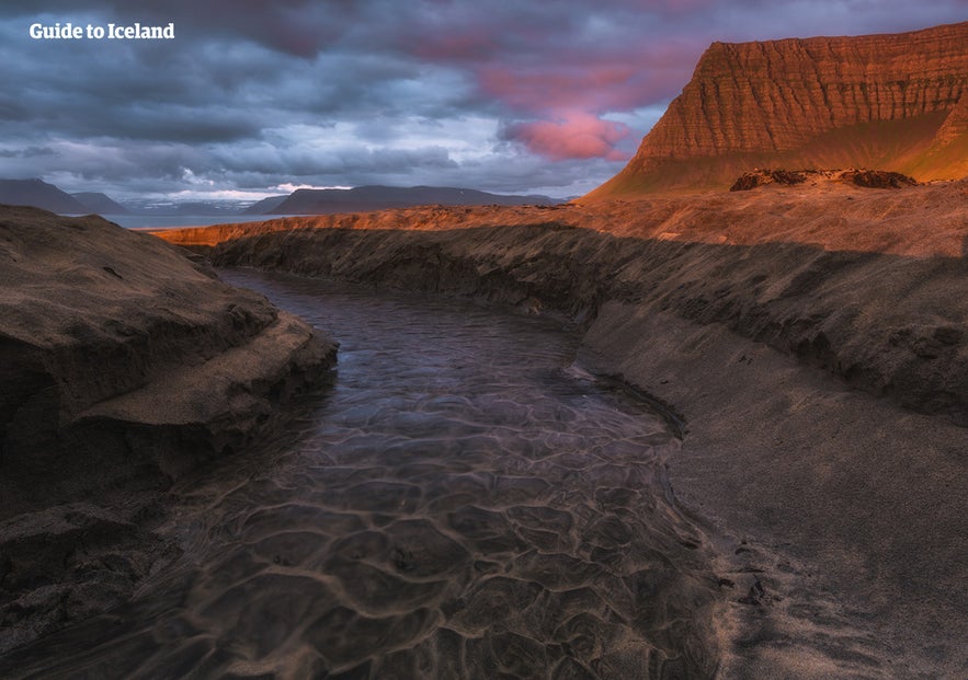 Sunset over Laugardalsá River winding through dark sand with steep mountains glowing under dramatic skies in Iceland.