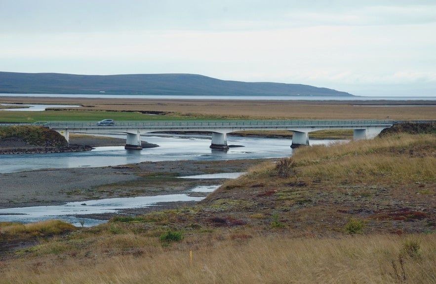 A bridge spans the calm waters of Midfjardara River in Iceland, with grassy plains and distant hills.