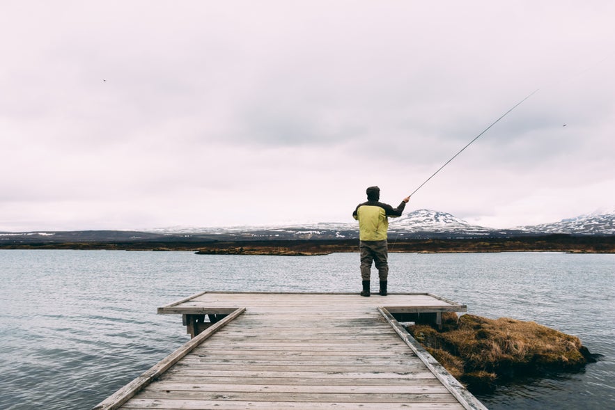 Man fishing from a wooden white pier on a lake at Thingvellir, Iceland, under cloudy weather.