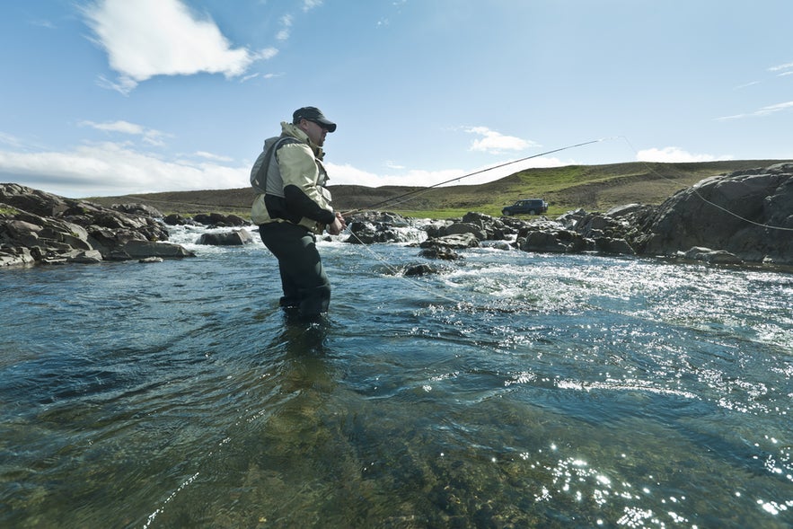 Man fly fishing in a clear Iceland river, wearing gear, with rocky hills and a vehicle in the distance under blue sky.