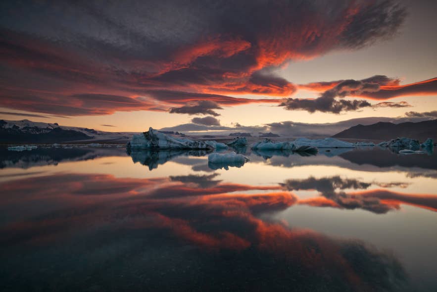 Solnedgang over Jökulsarlon-gletsjerlagunen i Island med isbjerge og farverige skyer, der spejler sig i det stille vand.