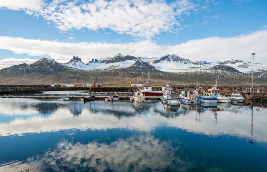 Fischerboote im ruhigen Hafen von Stodvarfjördur, Island, mit schneebedeckten Bergen, die sich im klaren Wasser spiegeln.