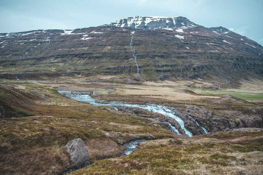 View of Seydisfjordur’s river and old power station with a snow-streaked mountain and cascading streams in the background.