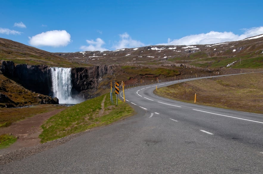 Gufufoss Waterfall near Seydisfjordur, Iceland viewed from Route 93 with a winding mountain road and clear sky.