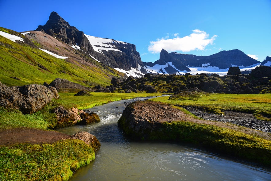 Storurd Valley in the Eastfjords with a winding stream, green moss, and snow-covered peaks under a clear blue sky.