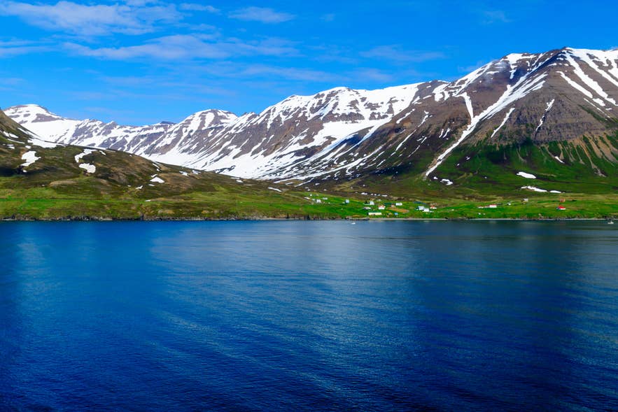 Le fjord Eyjafjordur aux eaux bleues calmes, avec quelques maisons et des montagnes striées de neige sous un ciel bleu éclatant.