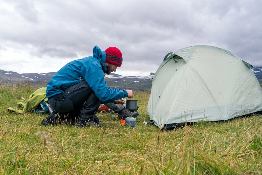 Person cooking by a tent while camping in Iceland, surrounded by grassy fields and distant snow-capped mountains.