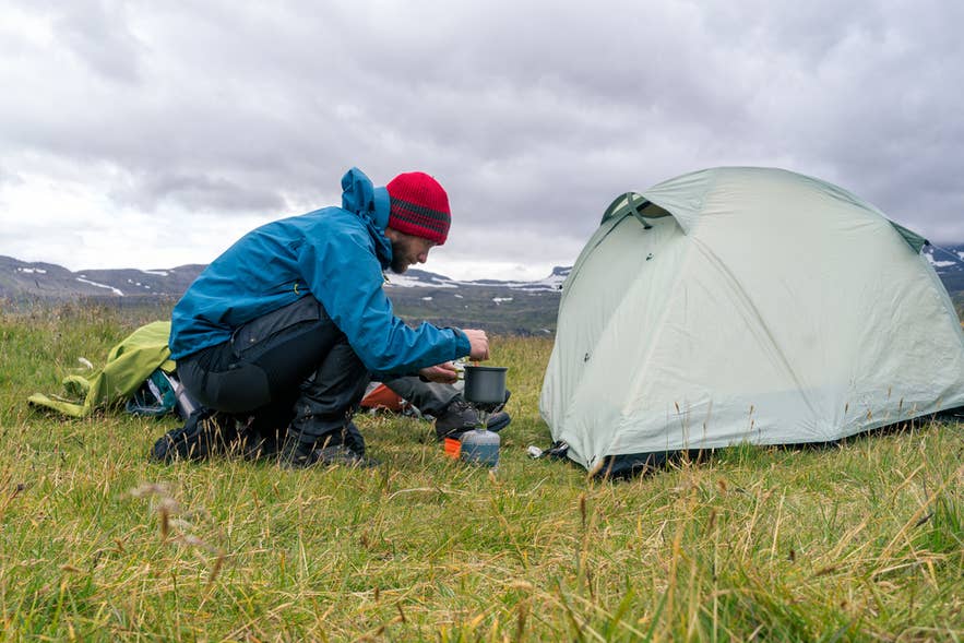 Person lagar mat vid ett tält under camping på Island, omgiven av gräsfält och avlägsna snötäckta berg.