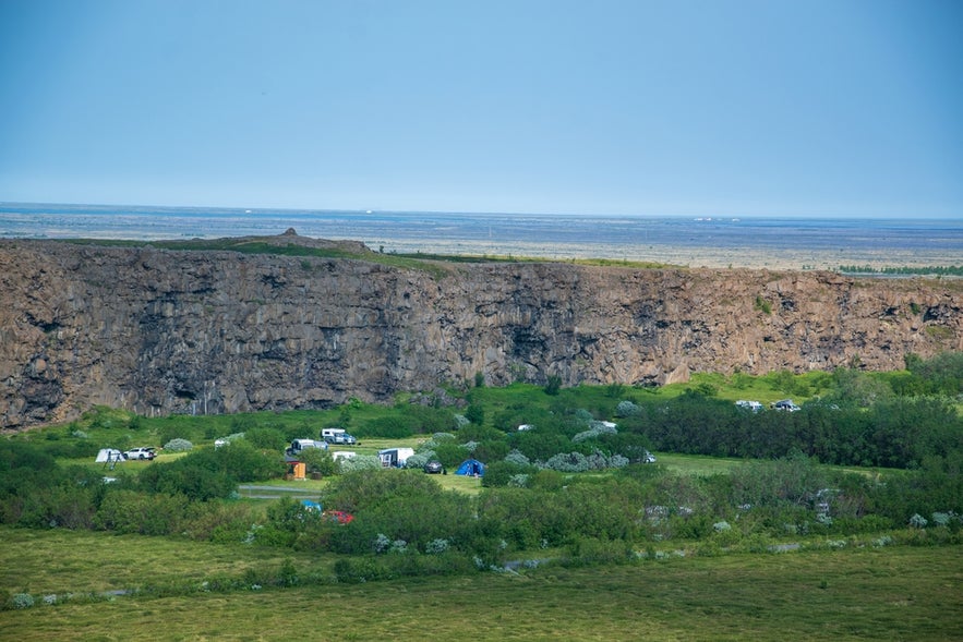 Campsite nestled in green vegetation below the towering cliffs of Asbyrgi canyon in Iceland, with tents and campers visible.