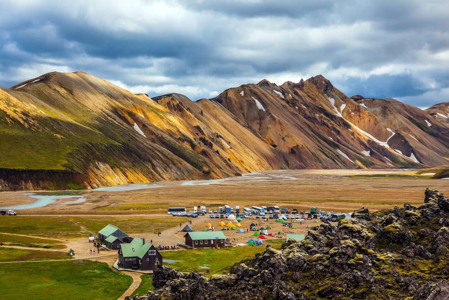 Landmannalaugar campingplats med färgglada tält, fordon och dramatiska, flerfärgade berg under molnig himmel.