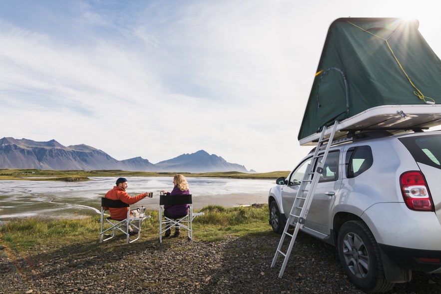 Couple camping in Hofn, Iceland, enjoying drinks by their car with rooftop tent and mountain views.