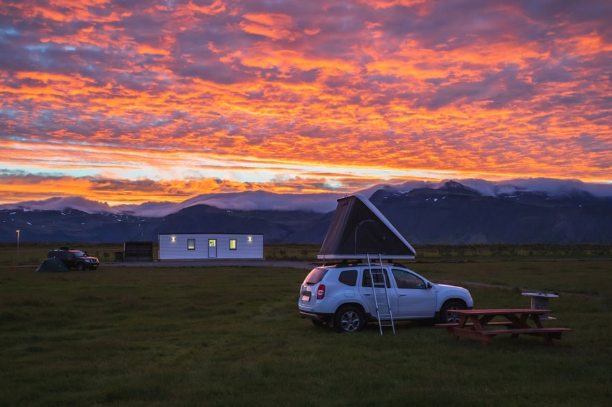 Camping in Iceland at sunset with a car rooftop tent, picnic table, and mountains in the background.
