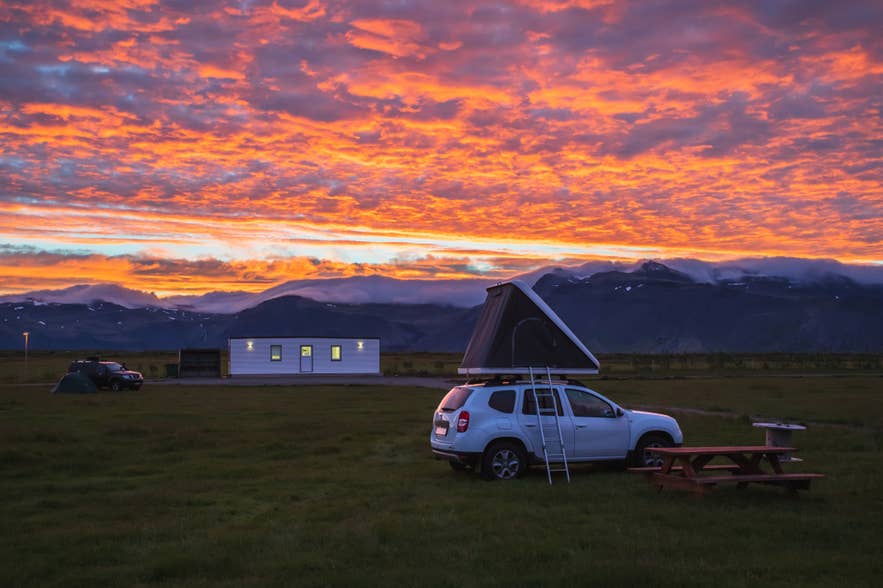 Camping på Island i solnedgången med bil och taktält, picknickbord och berg i bakgrunden.