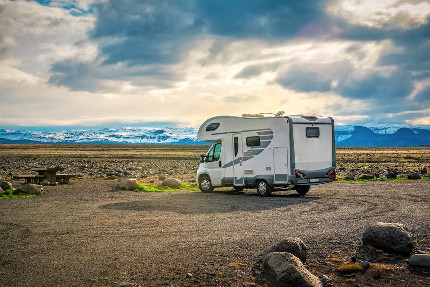 Camper van parked on rocky ground with picnic table, mountains, and cloudy sky—camping in Iceland.