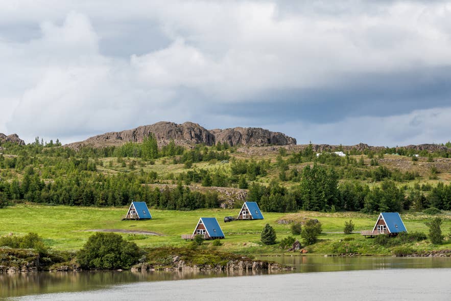 A-frame camping cabins with blue roofs near water in Egilsstadir, Iceland, surrounded by green hills.