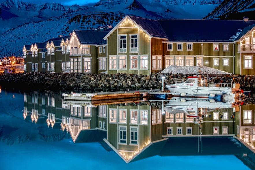 Colorful waterfront buildings and boats reflect on calm water at night in snowy Siglufjordur, Iceland.