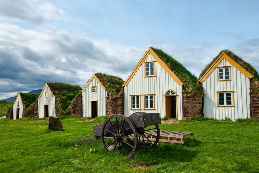 Traditional turf-roofed houses at Glaumbaer, Iceland, with a wooden cart on green grass under a cloudy sky.
