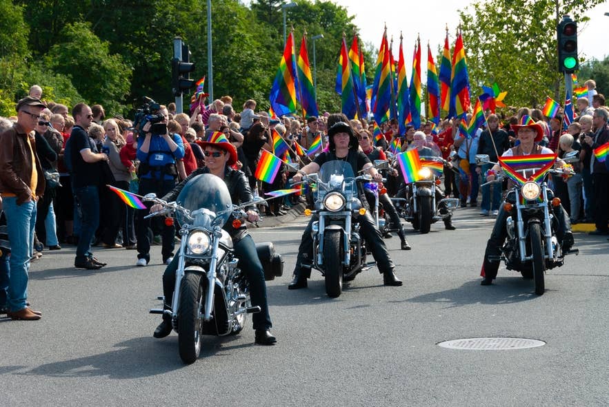 Motorradfahrer, die Regenbogenflaggen schwingen, führen eine Pride-Parade in Reykjavik an. Im Hintergrund ist eine Menschenmenge mit Fahnen zu sehen.