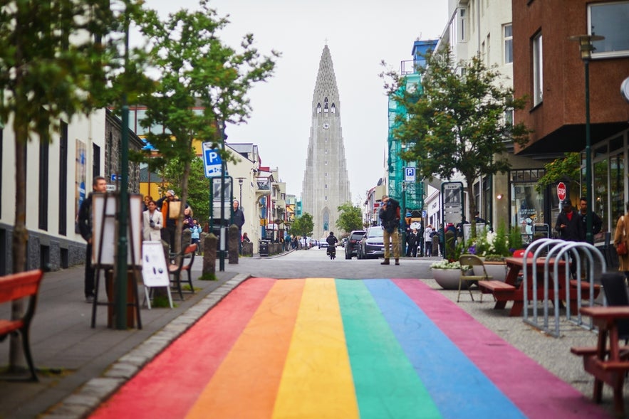 Rainbow-painted street in Reykjavik leads to Hallgrimskirkja church, with people walking and shops lining the road.