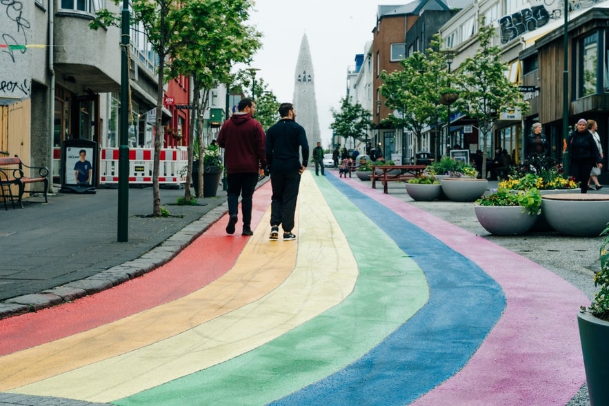 Two people walk on a rainbow-painted street in Reykjavík, Iceland, with Hallgrímskirkja church visible in the distance.
