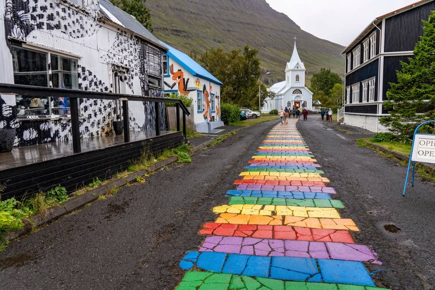 Rainbow-painted stone path leads to a white church in Seydisfjordur, Iceland, with colorful buildings on both sides.