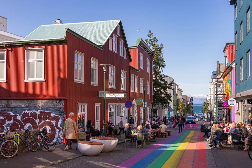 People enjoy a sunny day on Laugavegur Street in Reykjavík, with rainbow pavement and a view of the sea in the distance.
