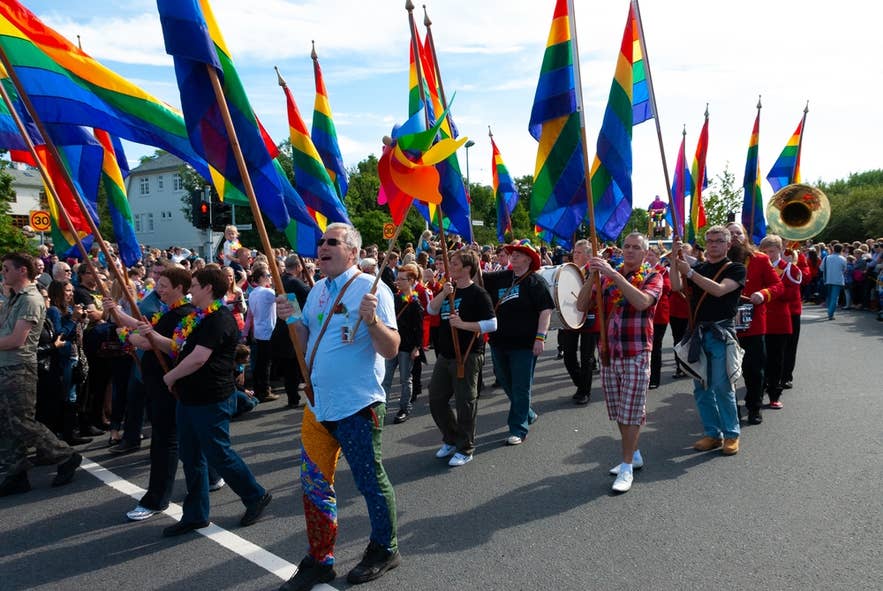 Menschen marschieren mit Regenbogenflaggen und einer Band während der Reykjavik Pride Parade an einem sonnigen Tag.