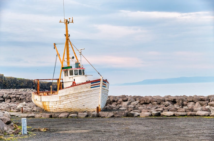 Weathered wooden fishing boat resting by the rocky seawall at Keflavik Harbor, with calm ocean views and a pastel sky in the background. Taking a walk along the harbor is one of the best things to do in Keflavik.