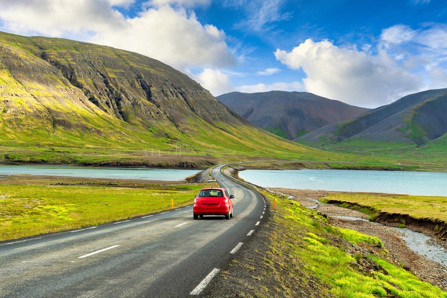 Red car driving through scenic countryside during summer in Iceland in June, with clear skies and green landscapes.