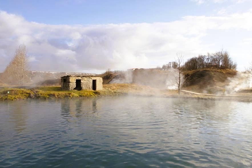 Dampf steigt aus der Secret Lagoon im Juni in Island auf, mit Steinhütte, klarem Wasser und grünen Hügeln im Hintergrund.