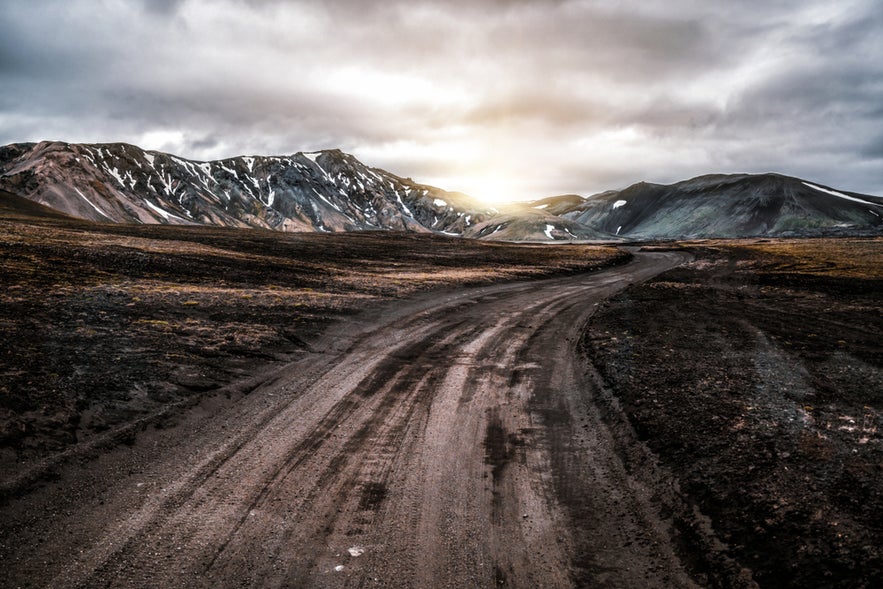 Gravel road winding through Icelandic Highlands at sunset, with rugged mountains and patches of snow in the distance.