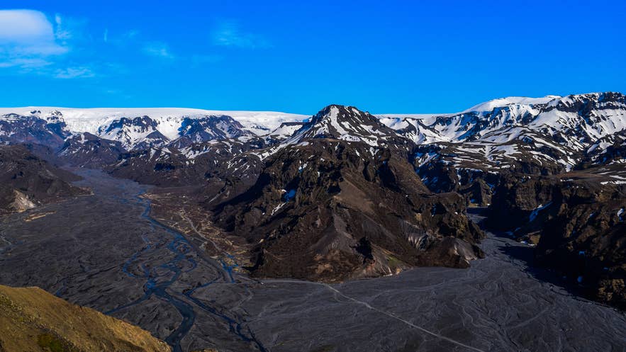 Luftfoto af Thorsmörk-dalen med forgrenede floder, mørkt vulkansk terræn og snedækkede bjerge under en klar blå himmel.