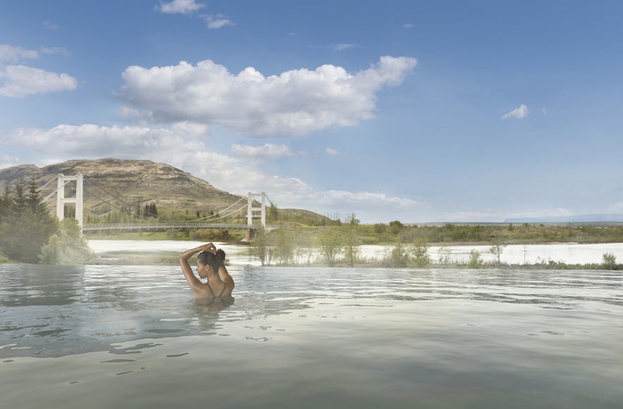 Woman relaxing in geothermal pool at Laugarás, opening in 2025, with views of a suspension bridge and Icelandic countryside.