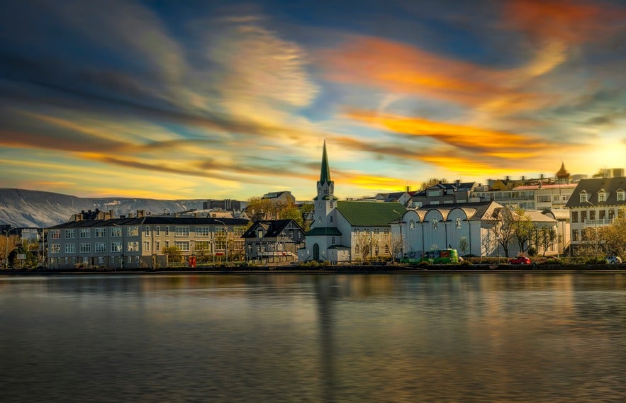 View of Reykjavík’s city center at sunset, featuring Fríkirkjan church and surrounding buildings reflected in the still waters of Tjörnin pond, under a dramatic, multicolored sky.