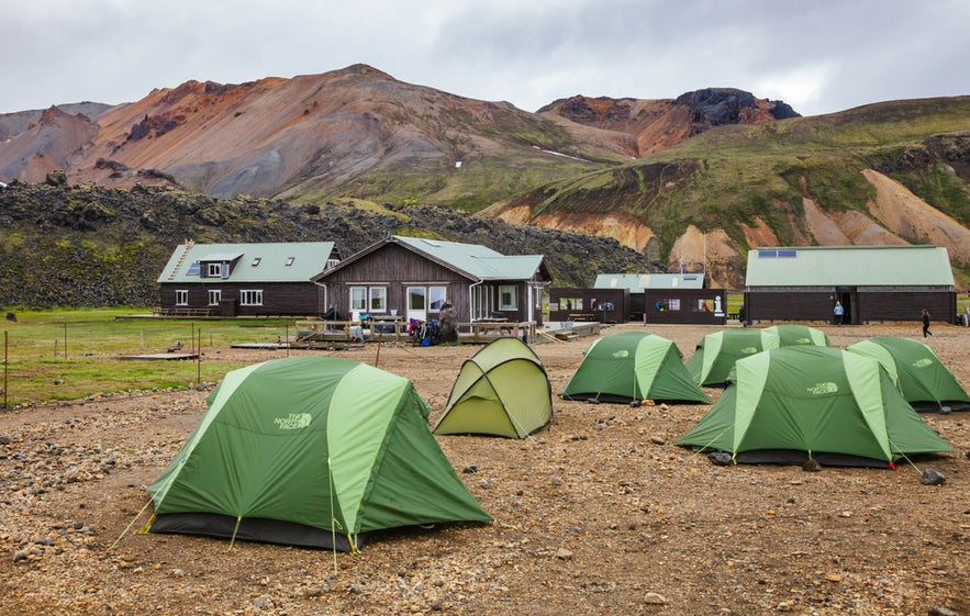 ChatGPT said: Green camping tents on rocky ground at Landmannalaugar, with colorful rhyolite mountains and huts in the Icelandic Highlands.
