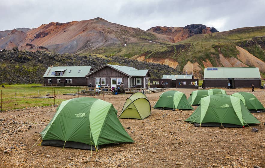 ChatGPT zei: Groene kampeertenten op rotsachtige grond bij Landmannalaugar, met kleurrijke ryolietbergen en hutten in de IJslandse Hooglanden.