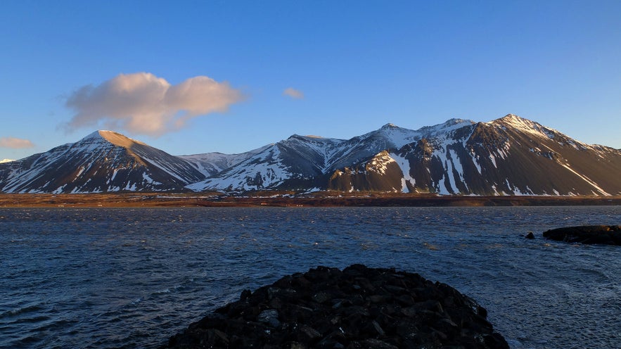 Snow-streaked Hafnarfjall mountain by the coast near Borgarnes, with evening light casting shadows over the water.