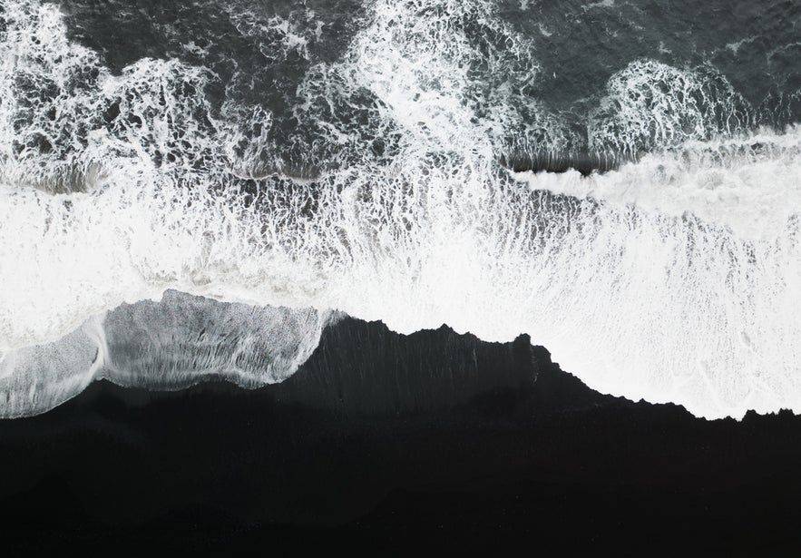 Aerial top view of ocean waves crashing onto the black sand of Solheimasandur Beach in Iceland.