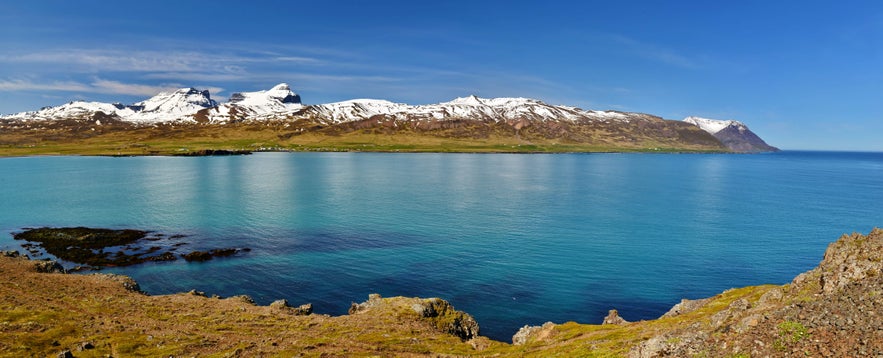 Maleriagtig udsigt over kysten n&aelig;r Borgarnes, Island, med turkisbl&aring;t vand, forrevne klipper og sned&aelig;kkede bjerge i det fjerne.