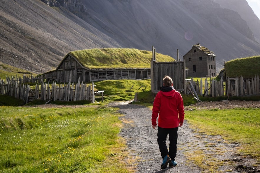 A man in a red jacket walks along a path toward the Viking Village film set, with grass-roofed buildings and the Vestrahorn Mountain in the background.
