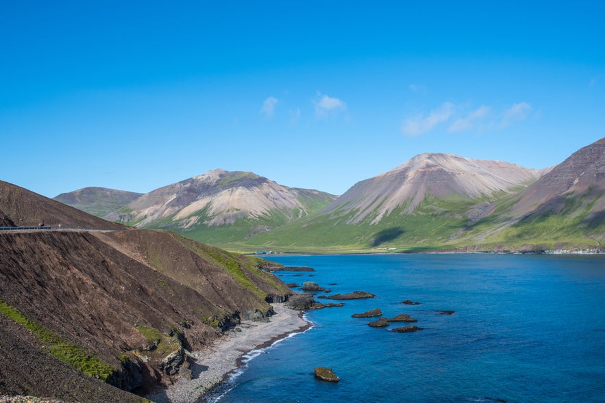 Njardvik Bay on the Reykjanes Peninsula, featuring a winding coastal road, rocky shore, and mountains under a clear blue sky.