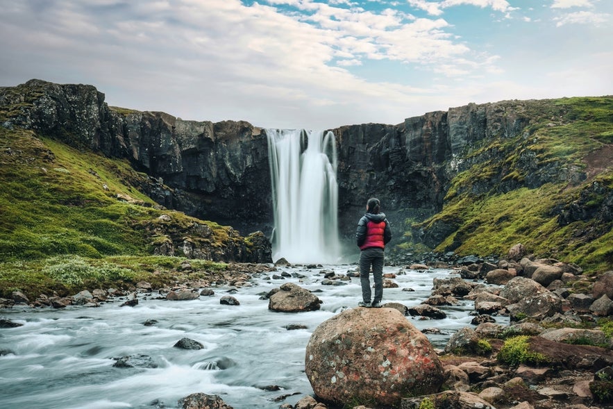 Person in red jacket stands on rock facing Gufufoss waterfall in Iceland's Eastfjords, surrounded by cliffs and river.