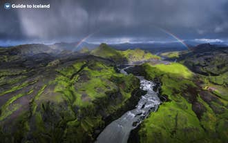 Un canyon de rivière et des montagnes verdoyantes dans les Hautes Terres islandaises, surmontés d’un arc-en-ciel.