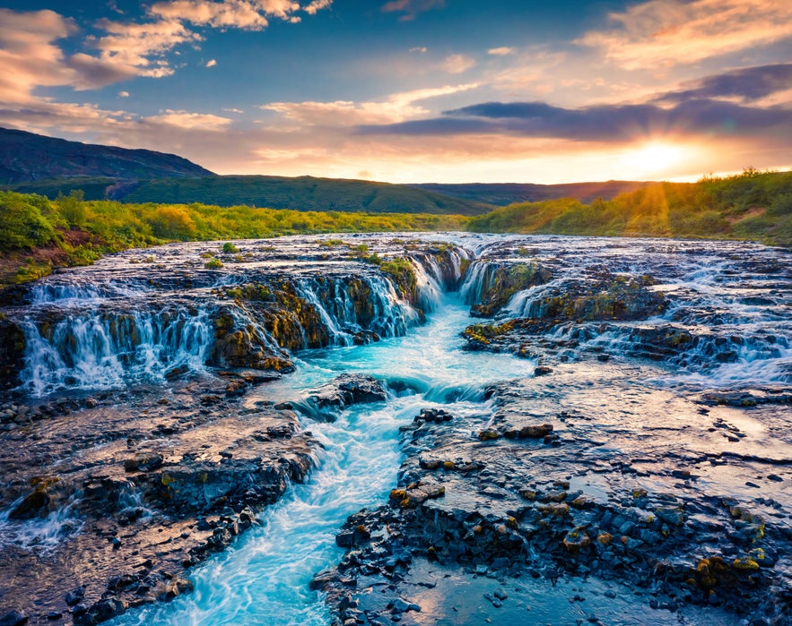 Bruarfoss waterfall in Iceland at sunset, with turquoise glacial water flowing over dark volcanic rock and lush greenery.