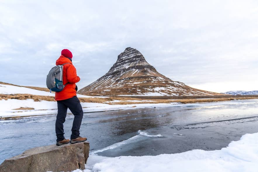 Randonneur en hiver à Kirkjufell portant des bottes, des couches chaudes et un bonnet rouge, illustrant quoi porter en Islande par temps neigeux.