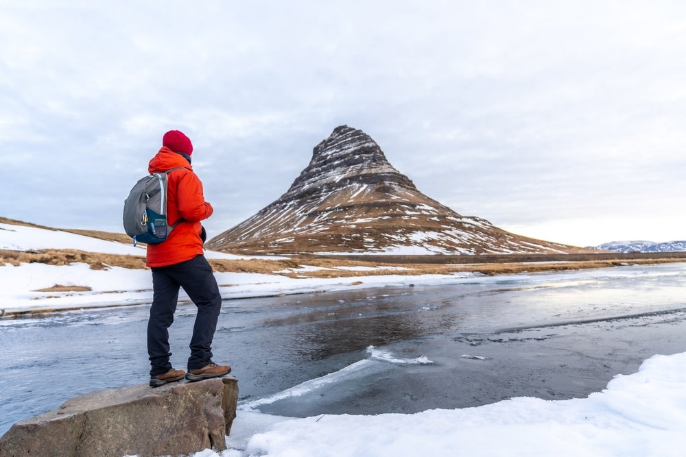 Winterwanderer am Kirkjufell mit Stiefeln, warmen Schichten und roter Mütze – so kleidet man sich in Island bei Schnee.